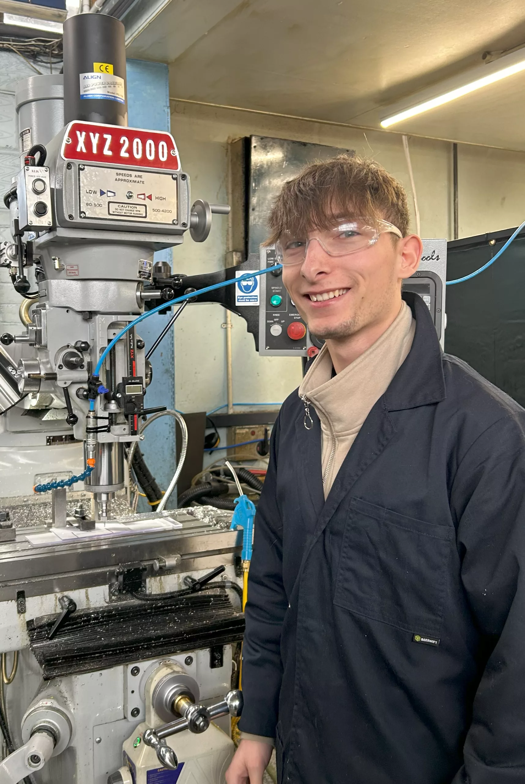 A young machining technician stands in front of an XYZ milling machine.